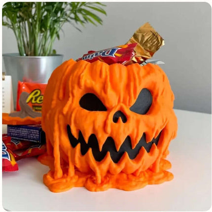 Halloween Melting Pumpkin Candy Bowl With Lid filled with assorted candies on a table with green foliage in the background.