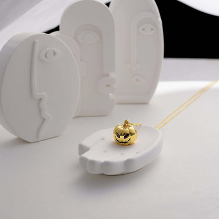 Womens Gold Pumpkin Diamond Halloween Necklace displayed on a white plate with minimalist art sculptures in the background.